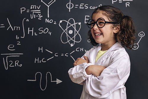 Little girl with brown hair and glasses in front of a blackboard