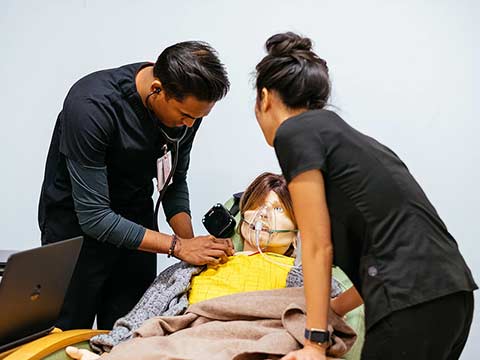 Two nurses practice procedures on a mannequin patient sitting up in a bed.