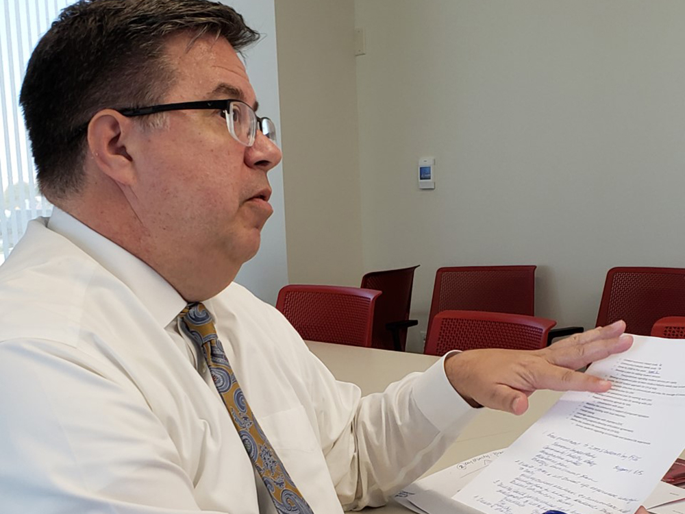 A dark haired man wearing glasses pointing to a piece of paper with notes