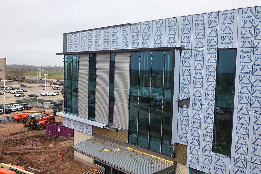 Photo of a side of building under construction. The exterior walls are covered in sheetrock. Construction equipment and materials are sprawled along the dirt ground.