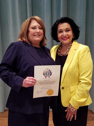 A blonde woman holding a certificate standing next to a dark-haired woman in a yellow jacket