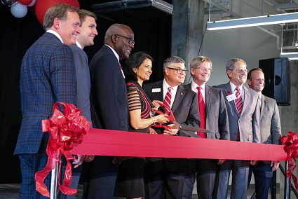 A group of men in suits stand around a man and woman holding a large open pair of scissors over a large red ribbon