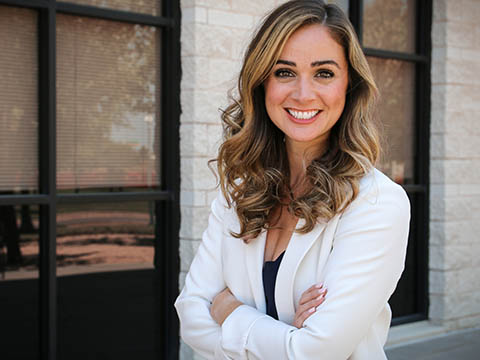 Portrait of a light brown-haired woman in a white business jacket standing in front of a cream brick wall with tall windows