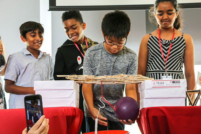 A young student hangs a weighted ball from a small flat bridge made of craft sticks to test the structure. He is flanked by his teammates.