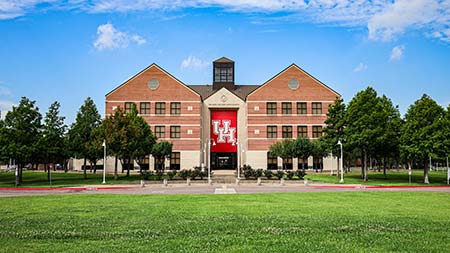 Photo of the Albert & Mamie George Building. On the front of the three story brick building is a large red UH banner. Green trees surround the building.