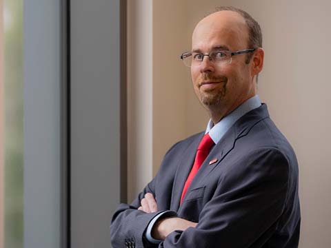 Portrait of a man with glasses and a beard standind sideways as he looks at the viewer.