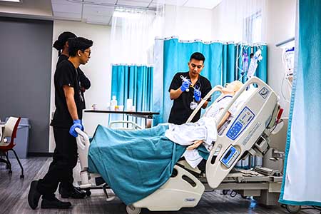 Two nursing students in black scrubs stand at the foot of a hospital bed. A third student stands at the side administering a syringe to a mannequin.