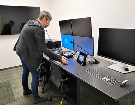 A woman stands over work station with 5 large monitors and flexible gooseneck microphones.