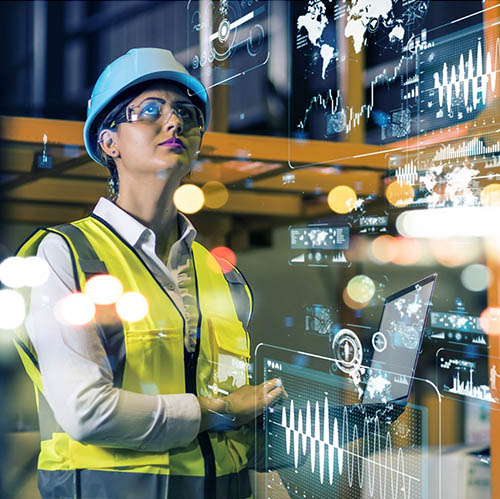 A women wearing a construction hardhat and safety vest inspects a construction area while holding a laptop.