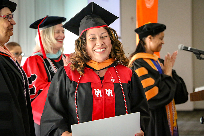 A student smiling in graduation regalia holding a diploma at a bed