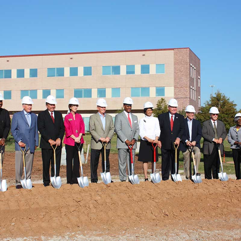 A ground of men and women in hardhats stand in a line holding shovels on a mound of dirt.